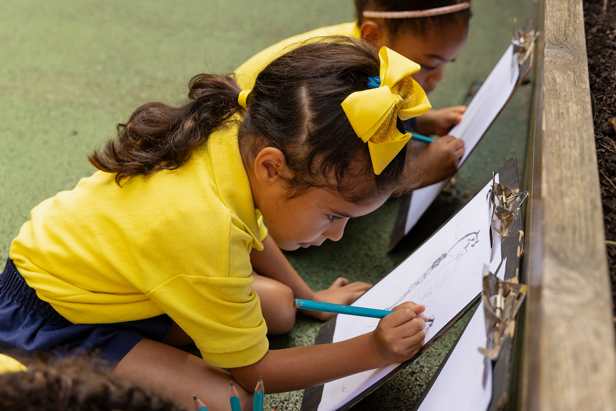 Child in a yellow shirt and bow draws on paper with a blue pencil, sitting on the ground next to another child.